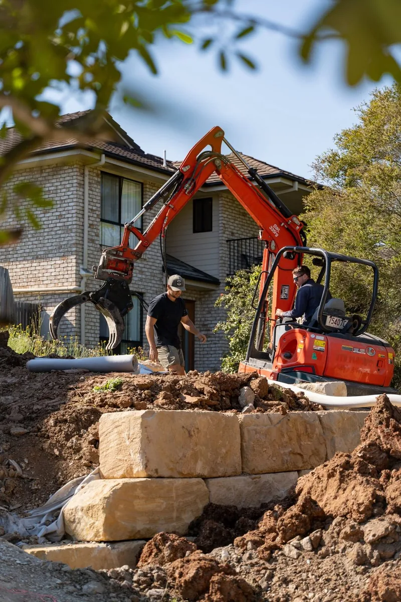 Excavator placing sandstone blocks on a residential retaining wall